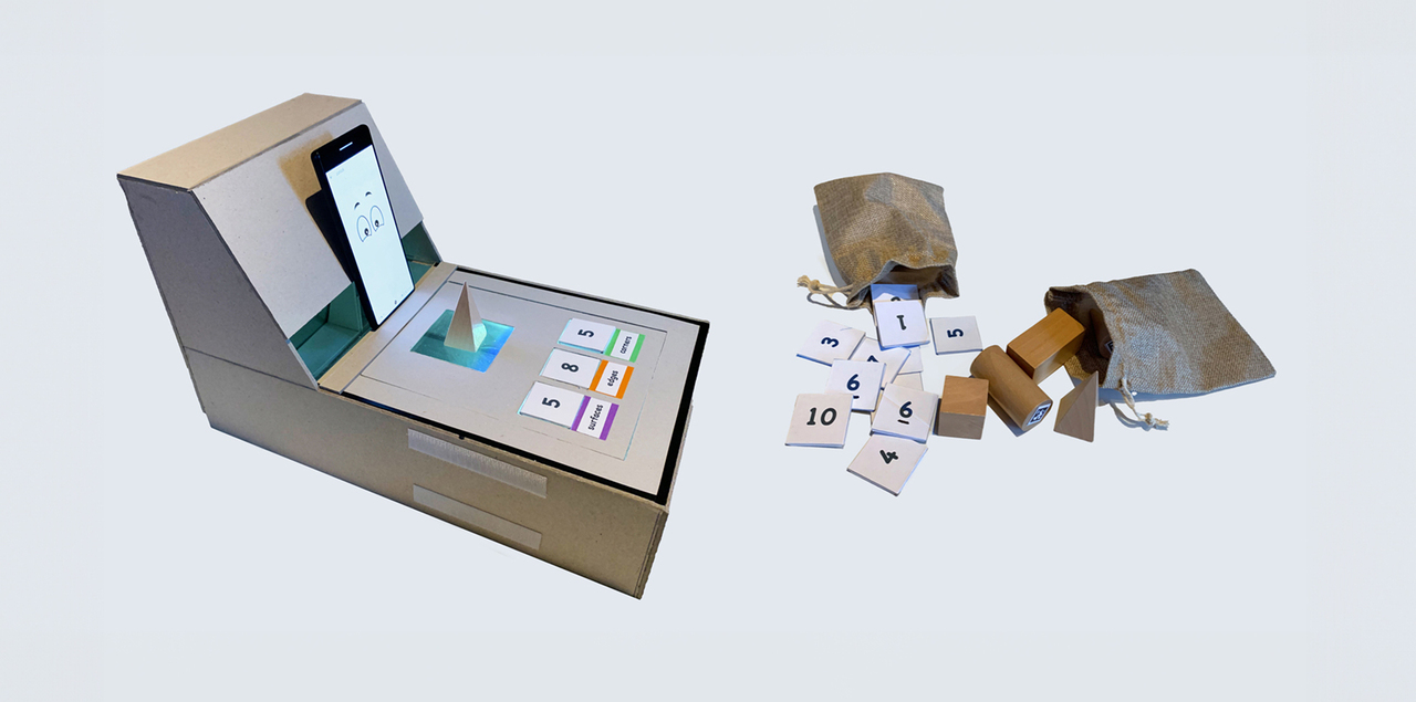 Cardboard Playmake prototype with a smartphone interface, number cards, and wooden geometric blocks spilled from small fabric bags.